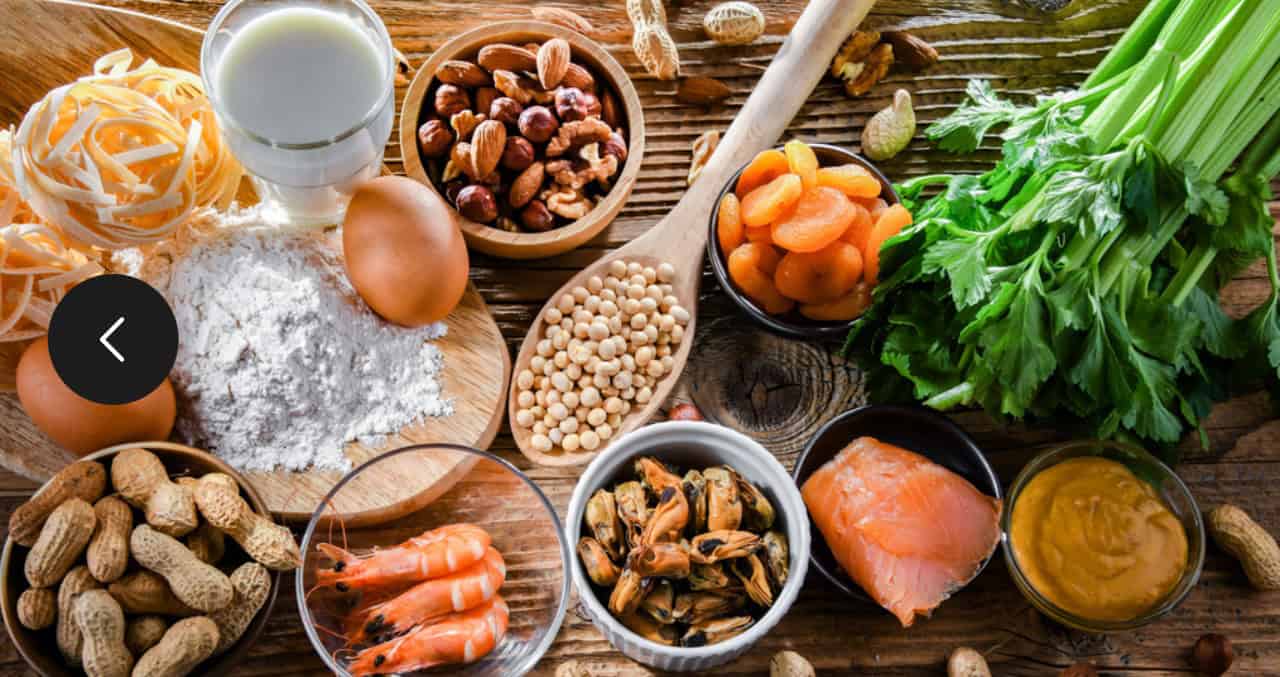 A wooden table displaying pasta, flour, eggs, almonds, beans, peanuts, apricots, celery, peanut butter, fish, and prawns.